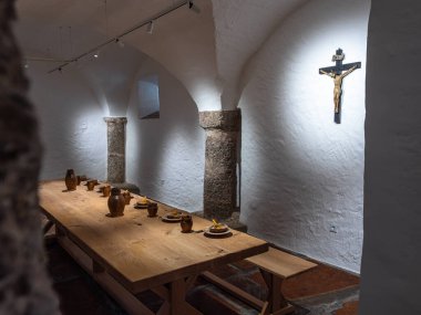 Michaelbeuern, Avusturya - 22 Ekim 2024: Kil tabakları ve sürahilerle dolu uzun ahşap bir masa ile manastır kırsal yemek odası. Çarmıh beyazlatılmış duvarda, tarihi bir ortam