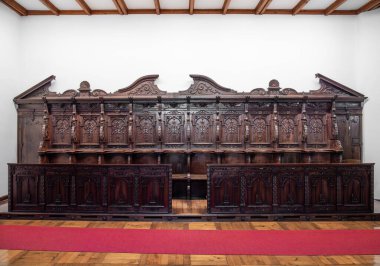 Michaelbeuern, Avusturya - 22 Ekim 2024: Büyük, karmaşık bir şekilde oyulmuş ahşap koro tezgahı, dekoratif detaylar, beyaz bir duvara dayanıyor. Birden fazla koltuk ve dekoratif paneller, vitrin işçiliği..