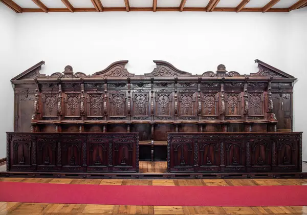 Michaelbeuern, Avusturya - 22 Ekim 2024: Büyük, karmaşık bir şekilde oyulmuş ahşap koro tezgahı, dekoratif detaylar, beyaz bir duvara dayanıyor. Birden fazla koltuk ve dekoratif paneller, vitrin işçiliği..