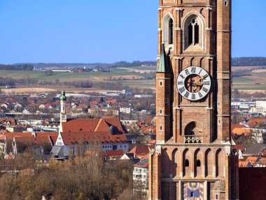 Landshut 'taki Aziz Martin Kilisesi' nin tarihi saat kulesi, büyük bir saat yüzüyle çevrili, kırmızı çatılı binaları ve arka planda yuvarlanan tepeleri olan küçük bir kasabanın manzarası..