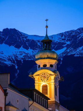 Günbatımında Innsbruck 'taki Kutsal Ruh' un hastane kilisesinin güzel aydınlatılmış saat kulesi karla kaplı dağların arkasına kurulmuş. Tarihin ve doğanın karışımı.