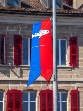 Nyon, İsviçre - 7 Mart 2025: Kırmızı panjurlu bir binanın önündeki direğe asılı mavi ve kırmızı ayrık desenli bir Nyon bayrağı.