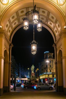 Zürih, İsviçre - 12 Aralık 2024: Süslü bir kemer yolu boyunca asılı fenerler ve bir heykelin yer aldığı bir Zürih caddesinin gece manzarası. Aydınlatılmış binalar ve bir otel tabelası görünüyor.