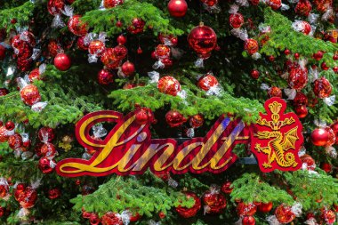 Zürih, İsviçre - 12 Aralık 2024: Kırmızı süslemelerle süslenmiş bir Noel ağacı, Lindt çikolata topları ve markalaşma, canlı bir tatil atmosferi sergiliyor.