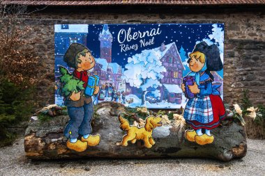 Obernai, Fransa - 12 Aralık 2024: Obernai 'de karlı köy arkaplanlı, geleneksel giyinmiş çizgi film karakterlerinin yer aldığı neşeli bir Noel duvar resmi.