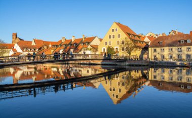 Landshut, Almanya - 19 Mart 2025: Isar 'ın sakin bir nehrine yansıyan renkli evlerle Landshut kasabası. Bir yaya köprüsü nehri geçiyor, üzerinde insanlar yürüyor..