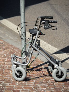 Metal bir direğe kilitlenmiş dört tekerlekli gri hareketli bir yürüteç kaldırıma park etmiş. Yürüteç, yaşlılar ve engelliler için tasarlanmış frenli bir koltuk ve gidona sahiptir..