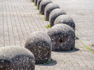 Asfaltlı bir yüzey üzerinde yuvarlak, yuvarlak taşlı bir sıra, dokulu ve yıpranmış bir görünüm sergiliyor. Sahne açık havada, yeşil alan kaldırıma bakıyor..
