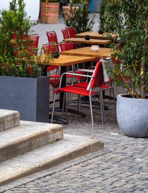 Etrafı yeşilliklerle çevrili ahşap masalar ve kırmızı sandalyelerle çevrili rahat bir kafeterya sahnesi. Çakıl taşı yolu ortama cazibe katıyor, rahatlama ya da sosyal toplanma için ideal..
