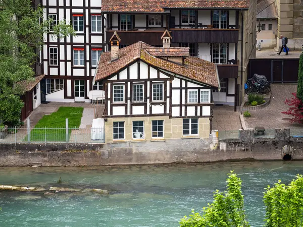 Bern, İsviçre - 8 Mayıs 2025: Bern 'deki Aare nehri kıyısındaki geleneksel yarı keresteli bir evin, yemyeşil ve daha büyük bir tarihi binayla çevrili bir manzarası.