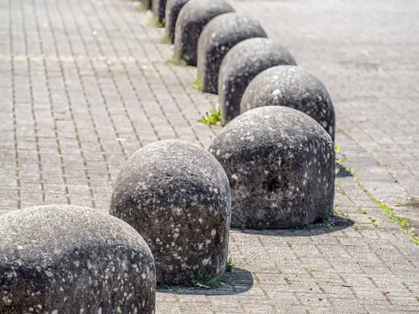 Asfaltlı bir yüzey üzerinde yuvarlak, yuvarlak taşlı bir sıra, dokulu ve yıpranmış bir görünüm sergiliyor. Sahne açık havada, yeşil alan kaldırıma bakıyor..