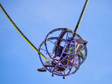 Havada asılı duran küresel bir kafesin olduğu heyecan verici bir lunapark gezisi, içinde iki binici, açık mavi bir gökyüzüne karşı.