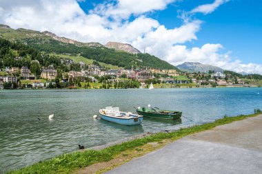 St. Moritz, İsviçre - 8 Temmuz 2024: Dağları, renkli binaları ve yemyeşil yeşillikleri olan Lakeside kasabası Sankt Moritz. Önde iki tekne var.