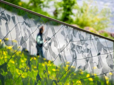 Bir yol boyunca yürüyen bulanık bir figür, yaprak desenli dekoratif bir ağ ekranına yansıyor. Arka planda canlı yeşil çimenler ve sarı çiçekler var, sakin bir açık hava manzarası yaratıyor..