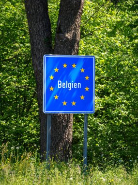 Belçika sınırını gösteren mavi bir yol işareti, yemyeşil ağaçlar ve yapraklarla çevrili. Tabelada, Avrupa Birliği 'ni temsil eden sarı yıldızlardan oluşan bir daireye sahip Belçika ismi yer alıyor..