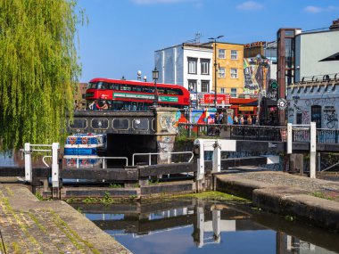 Londra, Birleşik Krallık - 28 Nisan 2025: Canlı bir Camden kasabası duvarları ve dükkanları olan bir Regent kanal köprüsünden geçen kırmızı çift katlı bir otobüsün yer aldığı canlı bir şehir manzarası.