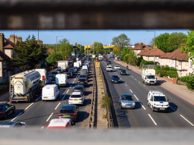 London, United Kingdom - April 28, 2025: Urban highway in London with heavy traffic, including cars and trucks, road is divided by a median, houses line the street. Viewed through a metal railing.