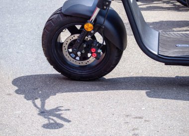Asfalttaki scooter tekerleğinin yakın çekimi, yerde lastik, fren sistemi ve gölgesi görünüyor..