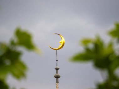 Bulanık yeşil yapraklarla çerçevelenmiş bir bina kulesinin tepesinde bulutlu bir gökyüzüne karşı, altın bir hilal..