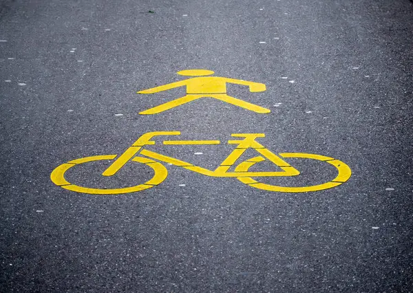 Sarı yaya ve bisiklet sembollerinin yer aldığı bir yol yüzeyinin yakın görüntüsü. Semboller asfalta boyanmış, yayalar ve bisikletçiler için belirlenmiş şeritleri gösteriyor..