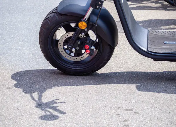 Asfalttaki scooter tekerleğinin yakın çekimi, yerde lastik, fren sistemi ve gölgesi görünüyor..