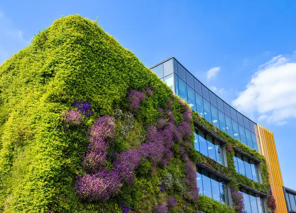 Açık mavi bir gökyüzünün altında yeşil bitkiler ve mor çiçeklerin yer aldığı, cephesini kaplayan yemyeşil dikey bahçeli modern bir bina..
