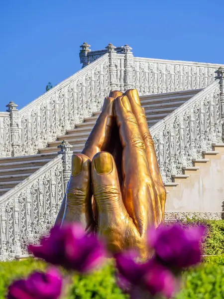 Londra, Birleşik Krallık - 28 Nisan 2025: Neasden Tapınağı bahçesinde dua eden ellerin altın heykeli, ön planda canlı çiçekler ve arka planda süslü merdivenler.