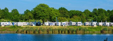 Maastricht, Netherlands - May 12, 2025: Panoramic view of a caravan park by a river Maas, featuring multiple caravans lined up along the shore, surrounded by lush greenery and trees.