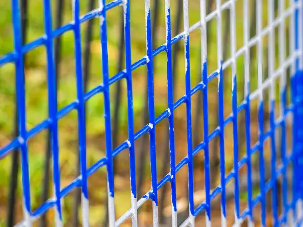 Mavi ve gri şebeke desenli renkli bir çitin yakın plan görüntüsü, bulanık doğal bir arkaplan karşısında ağın dokusunu ve yapısını gösteriyor..