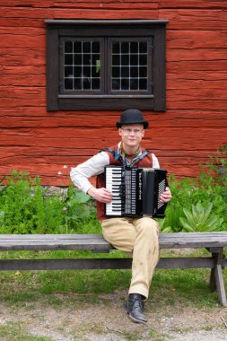 Sodertalje, İsveç - 6 Haziran 2025 - Kırsal bir kırmızı ahşap binanın önünde otururken akordeon çalan bir adam.