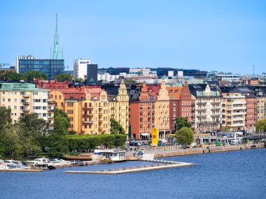 Stockholm, İsveç - 10 Haziran 2025: Stockholm 'de bir liman kenti boyunca manzaralı binalar, arka planda modern mimari ve açık mavi bir gökyüzü.