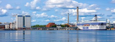 Stockholm, İsveç - 10 Haziran 2025: Büyük bir feribot, modern binalar ve kabarık bulutlu açık mavi gökyüzü içeren panoramik bir liman manzarası.