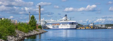 Stockholm, İsveç - 10 Haziran 2025: Büyük bir feribot, modern binalar ve kabarık bulutlu açık mavi gökyüzü içeren panoramik bir liman manzarası.