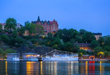 Stockholm, İsveç - 8 Haziran 2025: Alacakaranlıktaki bir rıhtım manzarası, bir tepenin üzerindeki tarihi bir otel binası, kıyıya kenetlenmiş HOTEL adlı bir tekne.