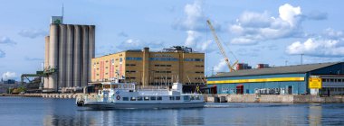 Stockholm, İsveç - 10 Haziran 2025: Büyük bir feribot, modern binalar ve kabarık bulutlu açık mavi gökyüzü içeren panoramik bir liman manzarası.