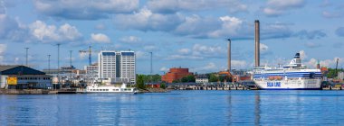 Stockholm, İsveç - 10 Haziran 2025: Mavi gökyüzü altında tüylü bulutlu büyük bir feribot, modern binalar ve endüstriyel yapıların yer aldığı manzaralı bir liman manzarası.