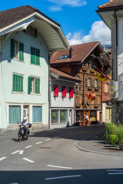 Interlaken, İsviçre - 17 Temmuz 2025: Geleneksel binalar, bayraklar ve açık mavi gökyüzü altında scooter süren bir kişi.