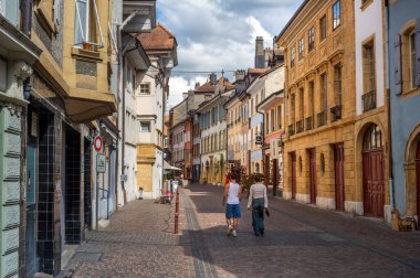Yverdon les Bains, İsviçre - 24 Temmuz 2025: Çekici bir Avrupa kasabasında renkli binalar, kaldırım taşları ve boş zamanlarında yürüyen iki kişinin yer aldığı pitoresk bir sokak manzarası.
