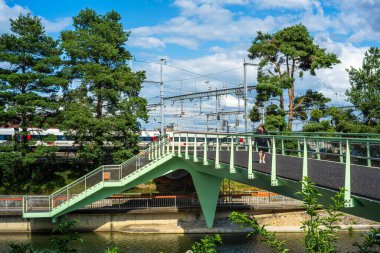Yverdon les Bains, İsviçre - 24 Temmuz 2025: Ağaçlarla çevrili, modern bir yeşil yaya köprüsü. Arka planda, tren rayları ve trenler görülebilir.