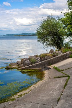 Sakin sular, kayalık kıyı şeridi ve yemyeşil verimli bir göl manzarası. Gökyüzü parçalı bulutlu, su yüzeyindeki yumuşak ışığı yansıtıyor..