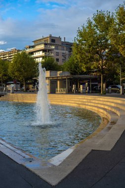 Lozan, İsviçre - 24 Eylül 2025: Mavi gökyüzü altındaki ağaçlar ve binalarla çevrili dairesel bir havuzda modern bir çeşme