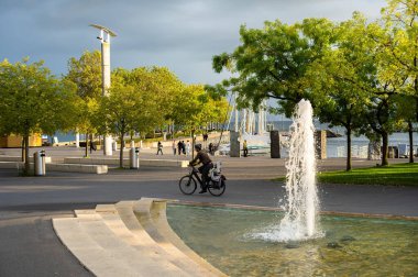 Lozan, İsviçre - 24 Eylül 2025: Bir bisikletçi, Lozan Ouchy 'un güneşli bir parkında ağaçlı ve deniz manzaralı bir çeşmenin yanından geçiyor.