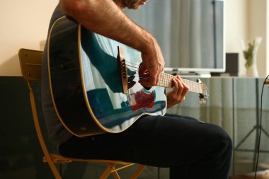 Evde, akustik gitarla parmaklarıyla akor çalan bir adamın ellerinin yakın çekimi.