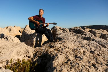 Koyu renk tişörtlü bir adam deniz kenarındaki kayalık bir alanda akustik gitar çalıyor.