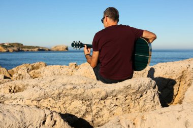 Koyu renk tişörtlü bir adam deniz kenarındaki kayalık bir alanda akustik gitar çalıyor.