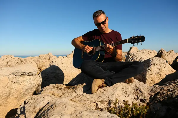 Koyu renk tişörtlü bir adam deniz kenarındaki kayalık bir alanda akustik gitar çalıyor.