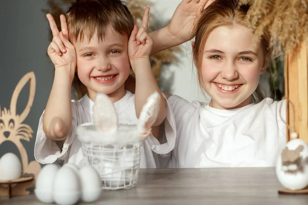 Christ is risen! Cheerful children boy and girl smile and show rabbit ears from their hands, symbols of the Easter holiday on the table