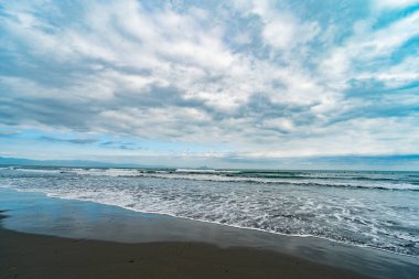 Dramatik bulutlu bir gökyüzünün altında yumuşak dalgalarla dolu engin kumlu bir sahil manzarası sunuyor.