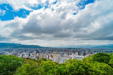 Canlı yeşil ağaçlar tarafından çerçevelenmiş geniş şehir manzarası. Kalabalık bir şehir üzerinde dramatik bir gökyüzü. Şehir panoraması.