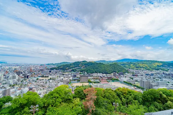 Arka planda dağlar olan panoramik şehir manzarası. Hareketli bir Japon şehrinin üzerinde dinamik bulutlar.